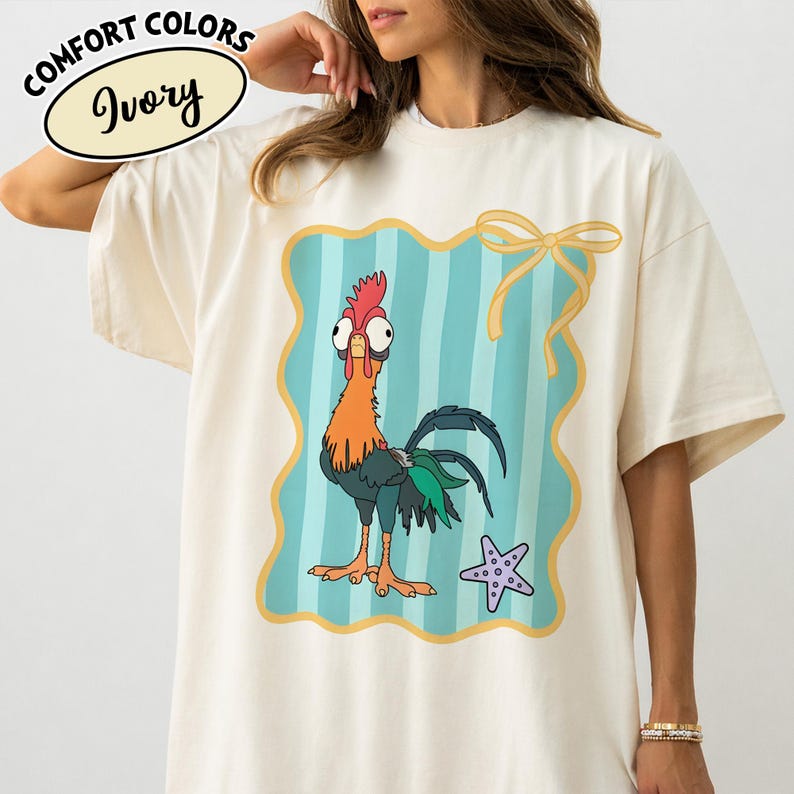 May include: Ivory-coloured t-shirt with a cartoon rooster design inside a teal and gold frame. The rooster has orange, red, and green details. The shirt also has the words "COMFORT COLORS" and "Ivory" printed on it.