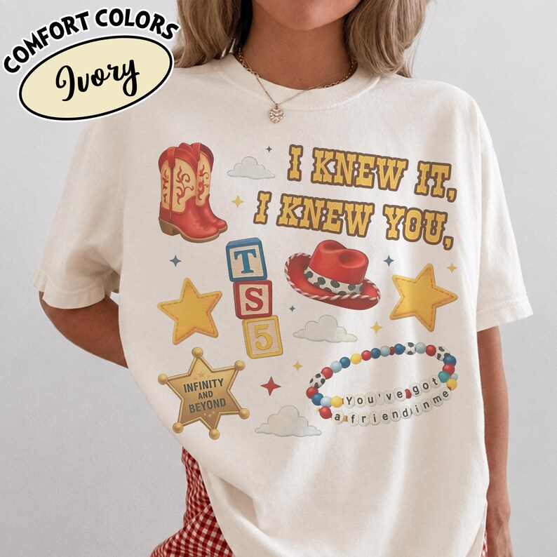 May include: An ivory t-shirt featuring a graphic print with a red cowboy boot, a red cowboy hat, yellow stars, a sheriff badge with the text INFINITY AND BEYOND, alphabet blocks reading TS 5, and a beaded bracelet with the text You've got a friend in me.