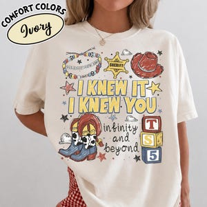 May include: An ivory t-shirt featuring a graphic print with the text "I KNEW IT I KNEW YOU", "infinity and beyond", and "TS 5". The design includes a sheriff badge, a cowboy hat, cow-print boots, a beaded bracelet, and colourful star accents.