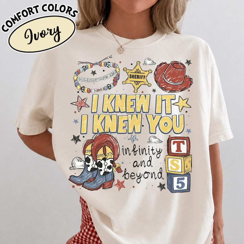 May include: An ivory t-shirt featuring a graphic print with the text "I KNEW IT I KNEW YOU", "infinity and beyond", and "TS 5". The design includes a sheriff badge, a cowboy hat, cow-print boots, a beaded bracelet, and colourful star accents.