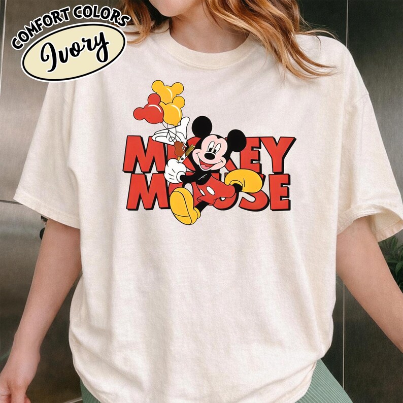 May include: Cream-coloured t-shirt with a Mickey Mouse graphic holding balloons. The design includes the words "Mickey Mouse" in red letters. The shirt has short sleeves and a crew neck. The words "Comfort Colors" and "Ivory" are printed on the top left.