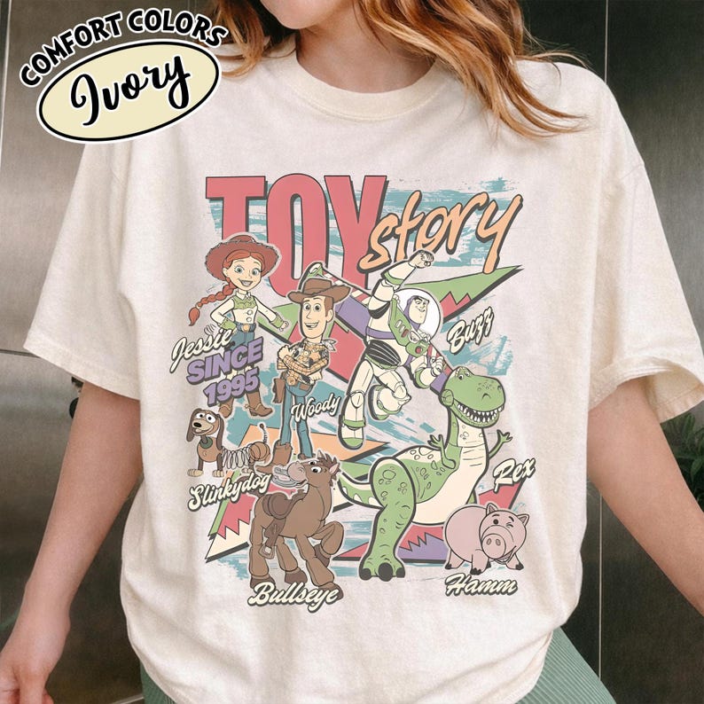 May include: Ivory-colored t-shirt featuring a colorful graphic of Toy Story characters, including Woody, Buzz Lightyear, and Rex. The design includes the text "Toy Story" and "Since 1995". The shirt has a crew neck and short sleeves.