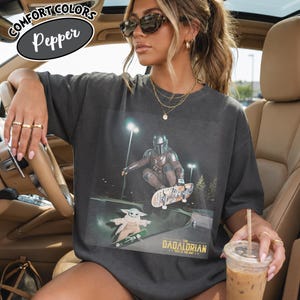 May include: Dark gray Comfort Colors Pepper t-shirt featuring a graphic of The Mandalorian and Grogu skateboarding. The design includes the text "The Dadalorian" and "This is the way". The shirt has a relaxed fit.