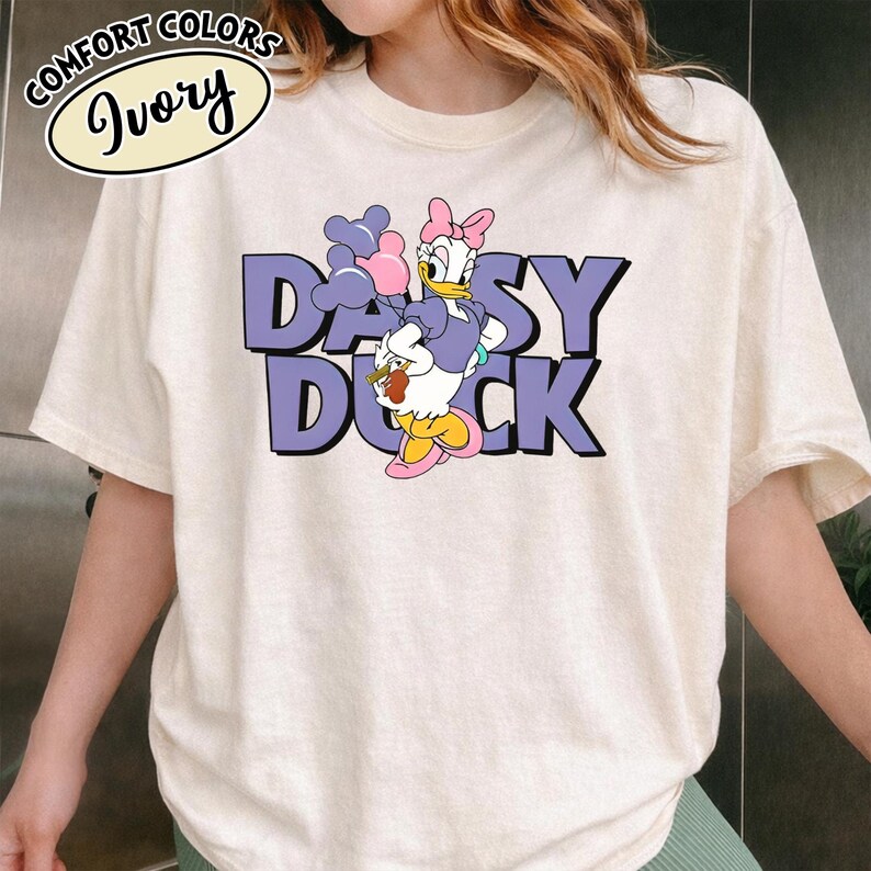 May include: Ivory t-shirt with a Daisy Duck graphic. The design features the text "DAISY DUCK" in purple, with Daisy Duck holding balloons and a snack. The shirt also has the words "COMFORT COLORS" and "Ivory" in a circular design.