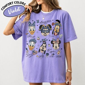 May include: A violet short-sleeve t-shirt featuring a graphic print of various cartoon characters including Mickey Mouse, Minnie Mouse, Donald Duck, Daisy Duck, and Goofy. The design includes stars, stripes, and the text 1776 - 2026.