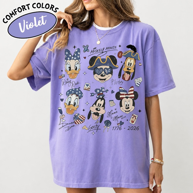 May include: A violet short-sleeve t-shirt featuring a graphic print of various cartoon characters including Mickey Mouse, Minnie Mouse, Donald Duck, Daisy Duck, and Goofy. The design includes stars, stripes, and the text 1776 - 2026.