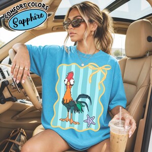 May include: A sapphire blue Comfort Colors t-shirt with a cartoon rooster design inside a striped frame. The rooster is orange, red, and green, with a purple starfish. The shirt has the Comfort Colors and Sapphire logos.