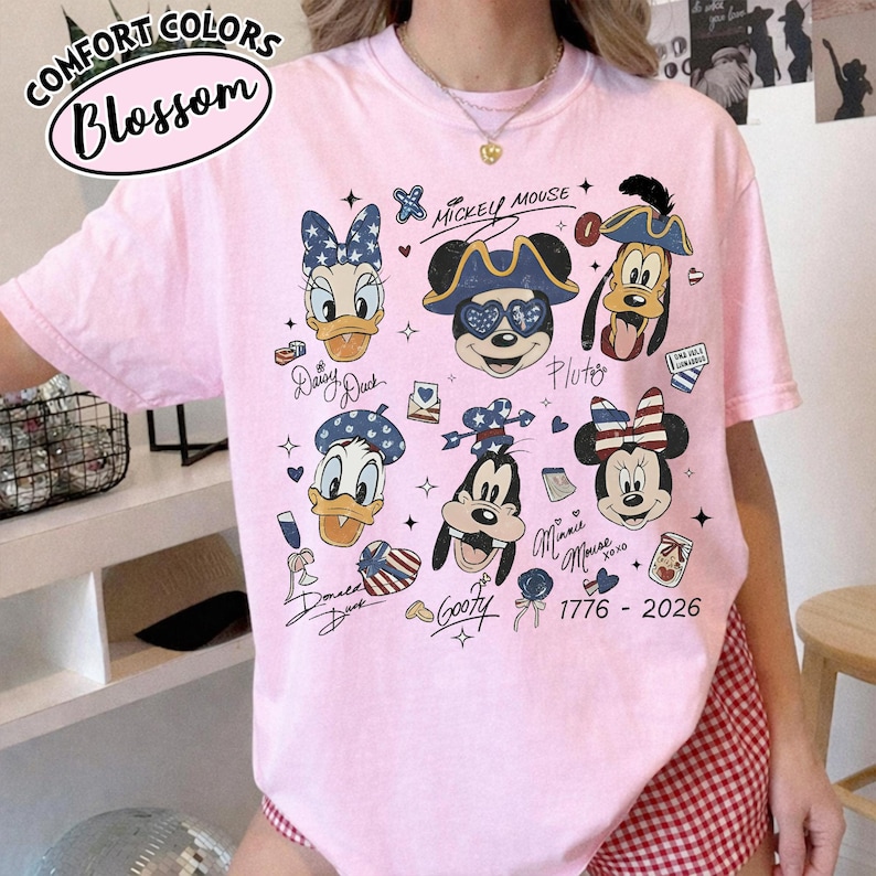May include: A light pink short-sleeve t-shirt featuring a graphic print of Disney characters including Mickey Mouse, Minnie Mouse, Donald Duck, Daisy Duck, Goofy, and Pluto. The design includes patriotic star and stripe patterns, text labels, and the date 1776-2026.