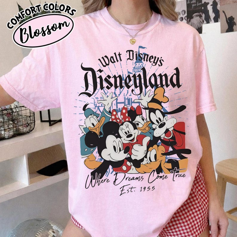 May include: A light pink t-shirt featuring the Disneyland logo and a graphic of Mickey Mouse, Minnie Mouse, Donald Duck, Daisy Duck, and Goofy. The shirt also has the text "Where Dreams Come True" and "Est. 1955". The top left corner has the text "Comfort Colors Blossom".