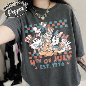 May include: A charcoal gray t-shirt featuring a graphic of cartoon characters wearing patriotic hats and holding an American flag. The design includes a checkered border and the text 4TH OF JULY EST. 1776. A label reads COMFORT COLORS Pepper.