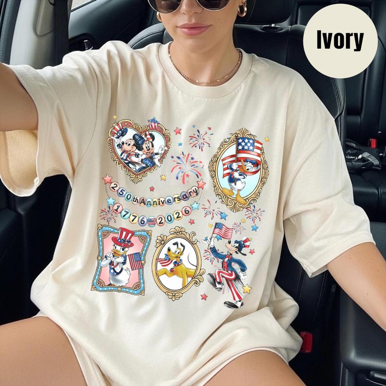 May include: Ivory-colored t-shirt featuring patriotic cartoon characters in ornate frames. The shirt has a design celebrating the 250th Anniversary, with fireworks and stars. The text "1776-2026" is also visible.