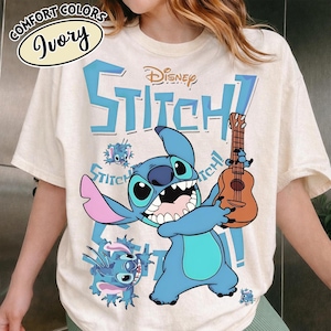 Pu&ograve; includere: T-shirt color avorio con un personaggio dei cartoni animati blu che tiene un ukulele. La maglietta ha la parola "Stitch" in grandi lettere blu, con il logo Disney sopra il personaggio. Le parole "Comfort Colors" e "Ivory" sono in un design circolare.
