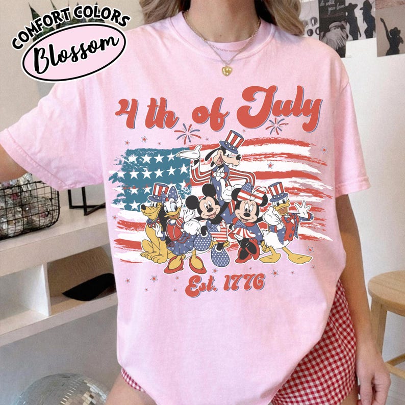 May include: Pink t-shirt with a graphic featuring Disney characters dressed in patriotic attire, including Mickey and Minnie Mouse, Goofy, and Donald Duck, in front of an American flag design. The text "4th of July" and "Est. 1776" are also displayed.