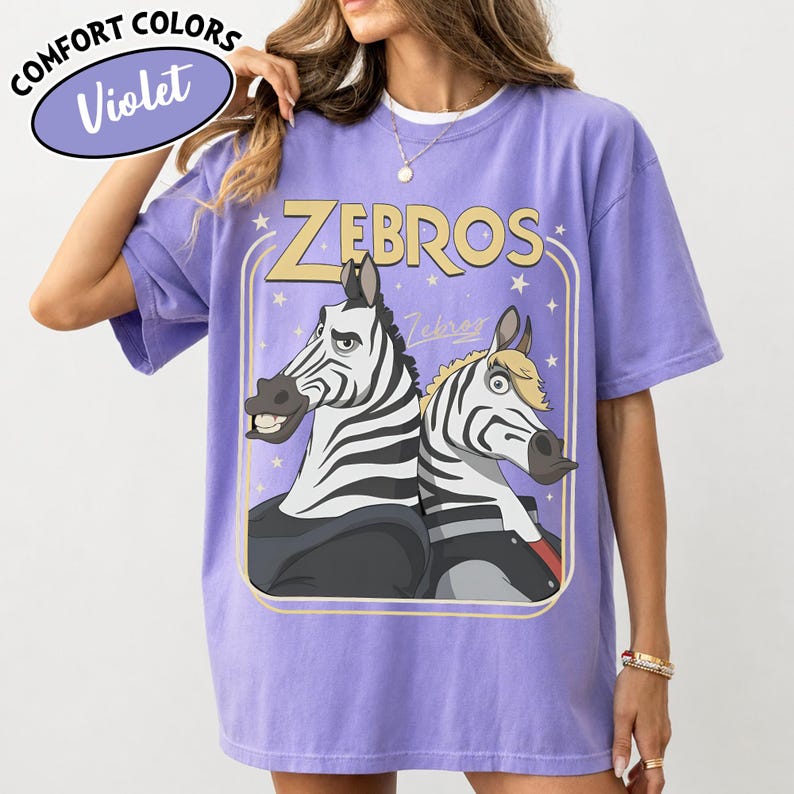Puede incluir: Camiseta violeta Comfort Colors con un dise&ntilde;o de dibujos animados de dos cebras y la palabra "ZEBROS" en una fuente retro. La camiseta tiene un corte holgado y mangas cortas. La camiseta es de color morado claro.
