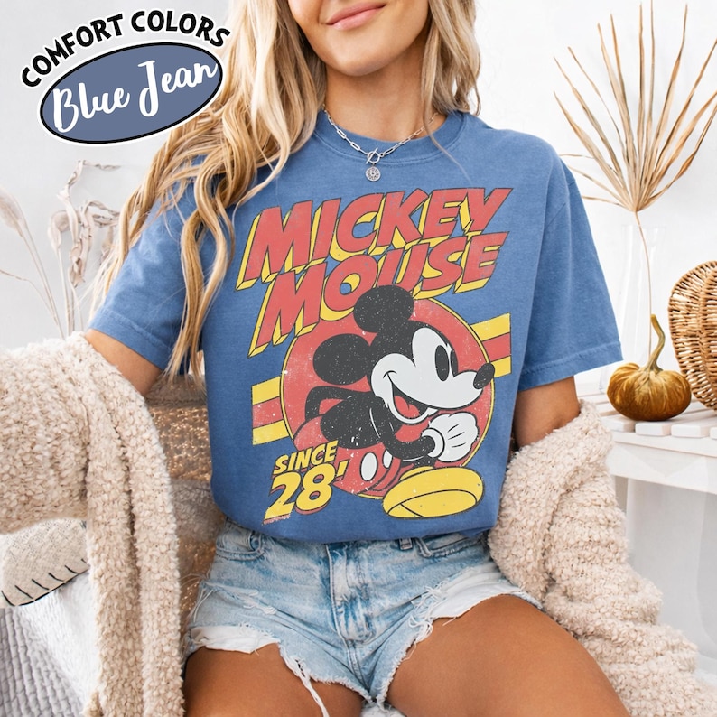以下が含まれることがあります： ヴィンテージミッキーマウスのデザインが施された青いTシャツ。デザインには、ミッキーマウスが笑顔で「Mickey Mouse」の文字が赤と黄色で描かれています。シャツには「Since 28」の文字も入っています。シャツには「Comfort Colors Blue Jean」と表示されています。