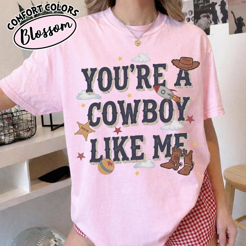 May include: A light pink short-sleeve t-shirt featuring the dark blue text "YOU'RE A COWBOY LIKE ME" centred on the chest. The graphic includes illustrations of a brown cowboy hat, a rocket ship, a sheriff star, a ball, and brown cowboy boots.