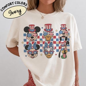 May include: Ivory-colored t-shirt featuring cartoon characters Mickey Mouse, Donald Duck, and Pluto, each wearing patriotic hats and sunglasses. The design includes a red, white, and blue checkered pattern and stars. The text "COMFORT COLORS Ivory" is at the top.