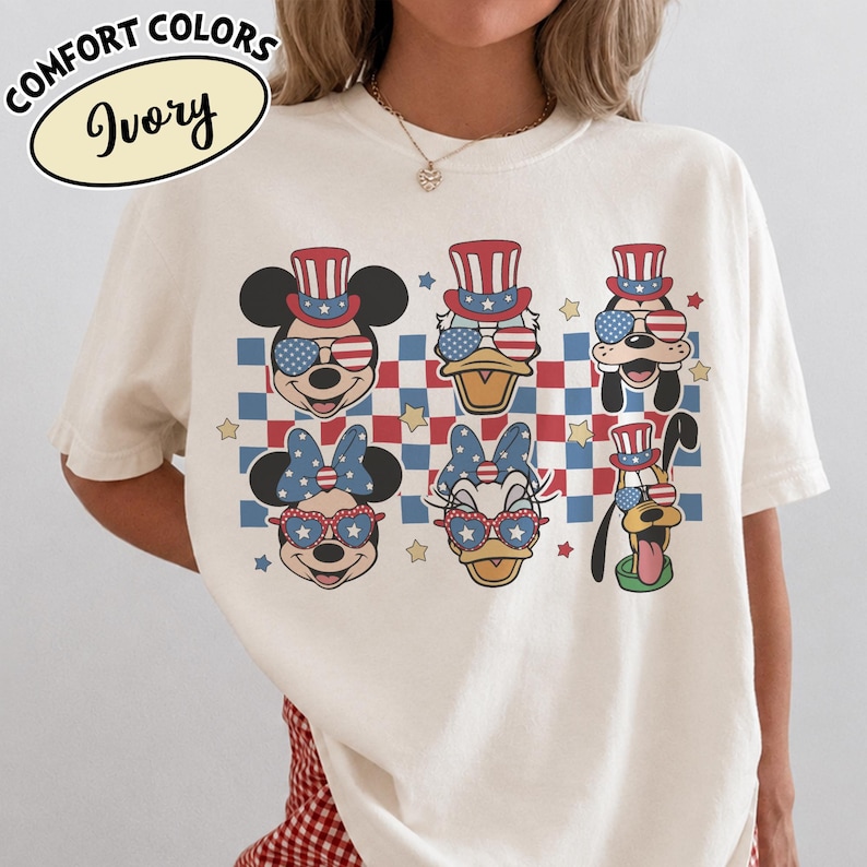 May include: Ivory-colored t-shirt featuring cartoon characters Mickey Mouse, Donald Duck, and Pluto, each wearing patriotic hats and sunglasses. The design includes a red, white, and blue checkered pattern and stars. The text "COMFORT COLORS Ivory" is at the top.