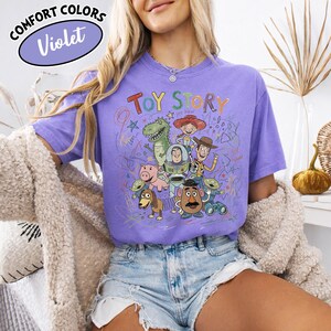 May include: A violet Comfort Colors t-shirt featuring a colorful graphic of Toy Story characters. The text "TOY STORY" is above the characters. The shirt has a crew neck and short sleeves.