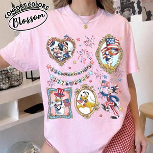 May include: A light pink t-shirt featuring framed cartoon characters, fireworks, and the text "250th Anniversary 1776-2026." The shirt has the words "Comfort Colors Blossom" in a circular design.