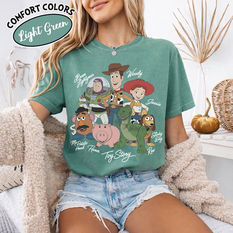May include: Light green t-shirt featuring a graphic of Toy Story characters, including Woody, Buzz Lightyear, and Jessie. The shirt has the words "Toy Story" printed at the bottom. The words "Comfort Colors" and "Light Green" are in the upper left corner.