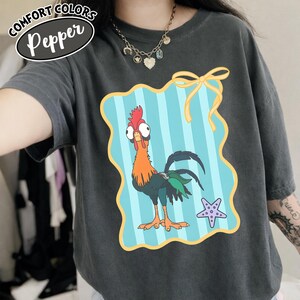 May include: Dark grey Comfort Colors t-shirt with a cartoon rooster. The rooster has a red comb, orange neck, and green tail feathers. It's set against a blue and white striped background with a yellow bow and a purple starfish.