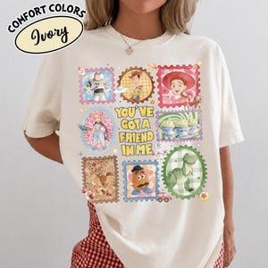 May include: Ivory-colored t-shirt featuring a colorful stamp design with characters from a popular animated movie. The stamps include images of a space ranger, a cowboy, a cowgirl, and other characters. The text "YOU'VE GOT A FRIEND IN ME" is also featured.