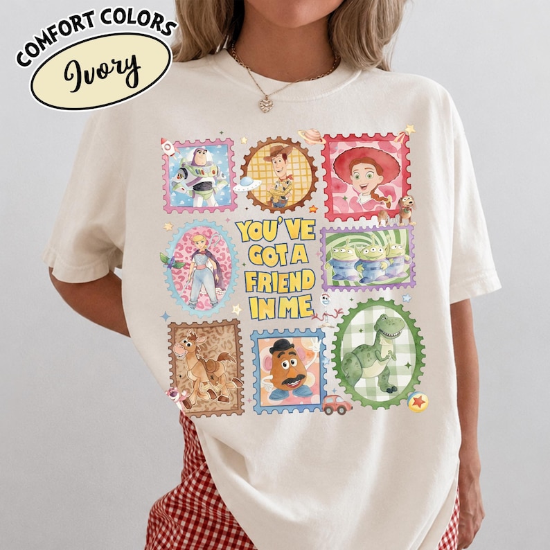May include: Ivory-colored t-shirt featuring a colorful stamp design with characters from a popular animated movie. The stamps include images of a space ranger, a cowboy, a cowgirl, and other characters. The text "YOU'VE GOT A FRIEND IN ME" is also featured.