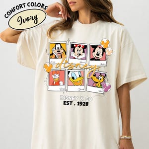 Op de afbeelding: Ivoor kleurig T-shirt met een Disney ontwerp. Het ontwerp bevat Disney-personages zoals Goofy, Mickey, Minnie, Donald, Daisy en Pluto. Het shirt heeft ook de tekst "Disney & Co. EST. 1928" en "Comfort Colors Ivory".
