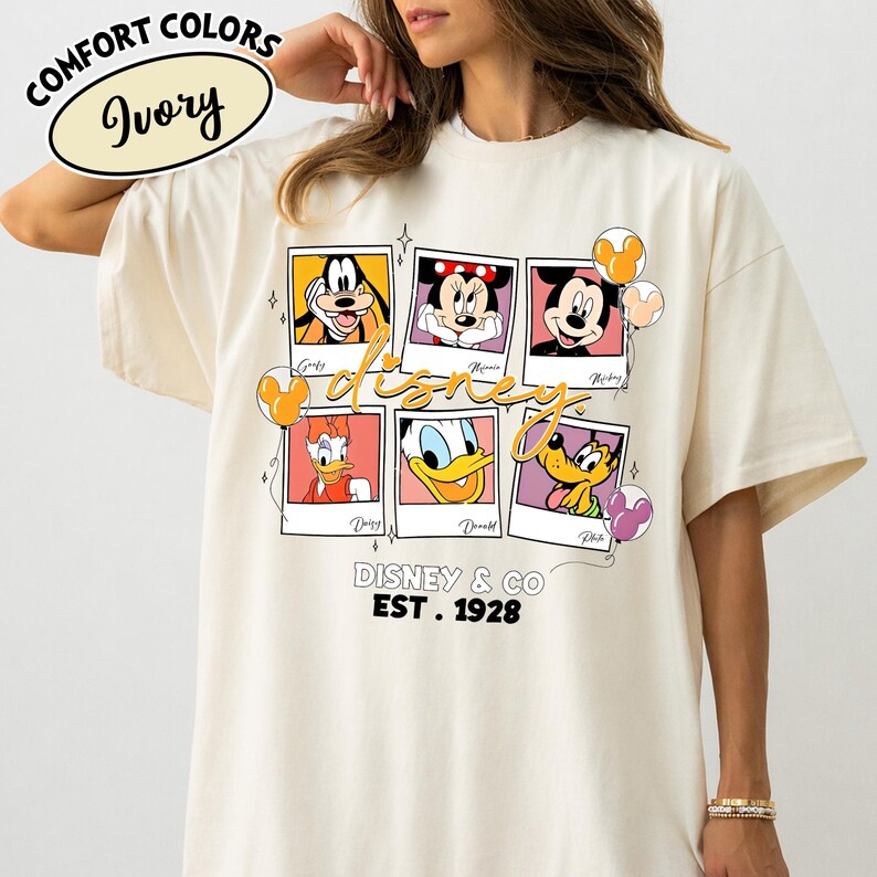Op de afbeelding: Ivoor kleurig T-shirt met een Disney ontwerp. Het ontwerp bevat Disney-personages zoals Goofy, Mickey, Minnie, Donald, Daisy en Pluto. Het shirt heeft ook de tekst "Disney & Co. EST. 1928" en "Comfort Colors Ivory".