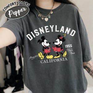 K&ouml;nnte beinhalten: Dunkelgraues T-Shirt mit einem Disneyland-Motiv mit Micky und Minnie Maus. Die Grafik enth&auml;lt den Text "Disneyland 1955 California" und "Mickey Mouse" und "Minnie Mouse". Das Shirt hat kurze &Auml;rmel und eine lockere Passform.