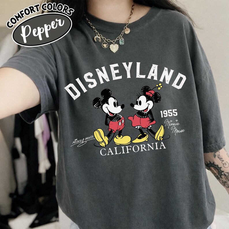 K&ouml;nnte beinhalten: Dunkelgraues T-Shirt mit einem Disneyland-Motiv mit Micky und Minnie Maus. Die Grafik enth&auml;lt den Text "Disneyland 1955 California" und "Mickey Mouse" und "Minnie Mouse". Das Shirt hat kurze &Auml;rmel und eine lockere Passform.