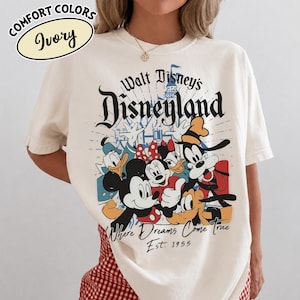 May include: Ivory-colored t-shirt featuring a graphic of Disney characters, including Mickey Mouse, Minnie Mouse, Donald Duck, and Goofy. The shirt has the text "Walt Disney's Disneyland" and "Where Dreams Come True Est. 1955". The shirt is a Comfort Colors brand.