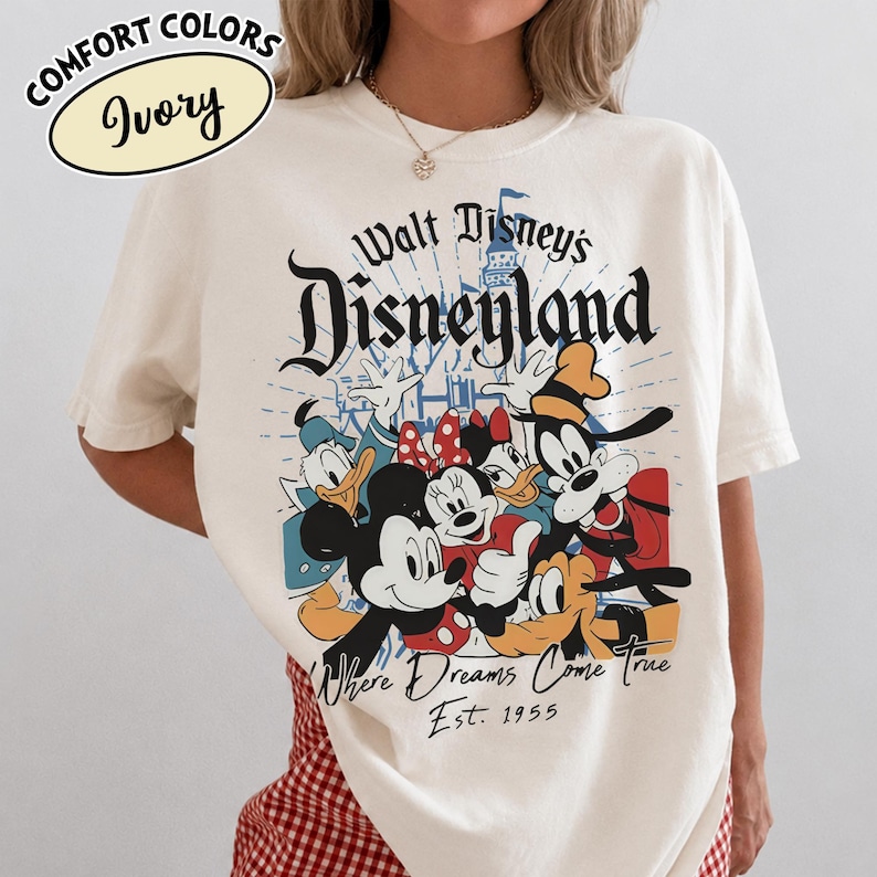 May include: Ivory-colored t-shirt featuring a graphic of Disney characters, including Mickey Mouse, Minnie Mouse, Donald Duck, and Goofy. The shirt has the text "Walt Disney's Disneyland" and "Where Dreams Come True Est. 1955". The shirt is a Comfort Colors brand.