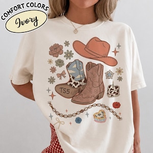 May include: An ivory t-shirt featuring a graphic print of a peach cowboy hat, two cowboy boots, flowers, a bow, a star, a cake, and a beaded string with the text I KNEW IT I KNEW YOU. One boot has a cow print and the text TS5 on the sole.
