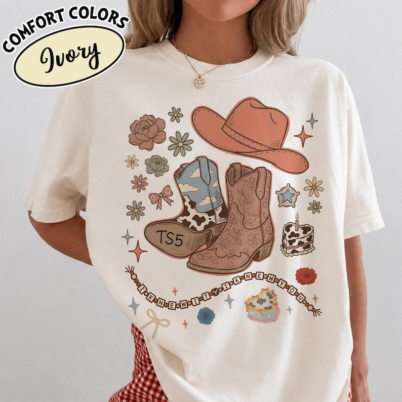May include: An ivory t-shirt featuring a graphic print of a peach cowboy hat, two cowboy boots, flowers, a bow, a star, a cake, and a beaded string with the text I KNEW IT I KNEW YOU. One boot has a cow print and the text TS5 on the sole.