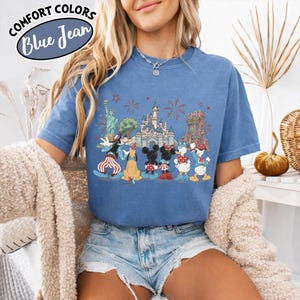 以下が含まれることがあります： 青色のComfort Colors Tシャツ。自由の女神や城など、象徴的なランドマークの前にディズニーのキャラクターが描かれたグラフィック。クルーネックと半袖。左上に「Comfort Colors Blue Jean」の文字。