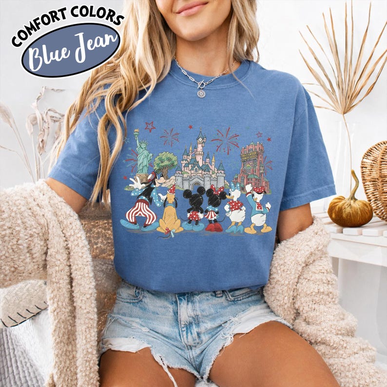 以下が含まれることがあります： 青色のComfort Colors Tシャツ。自由の女神や城など、象徴的なランドマークの前にディズニーのキャラクターが描かれたグラフィック。クルーネックと半袖。左上に「Comfort Colors Blue Jean」の文字。