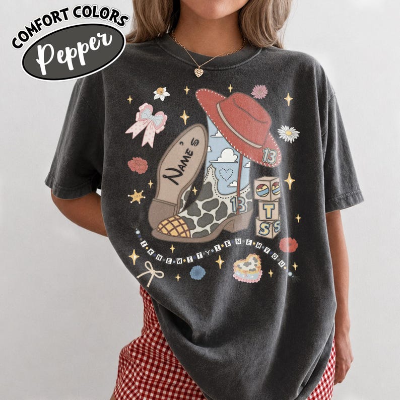 Puede incluir: Una camiseta gris carb&oacute;n de manga corta con un estampado gr&aacute;fico de una bota vaquera con manchas de vaca, un sombrero vaquero rojo, un lazo rosa y bloques de letras. El texto "NAME'S" y "I KNEW IT I KNEW YOU" est&aacute; impreso en la parte frontal.