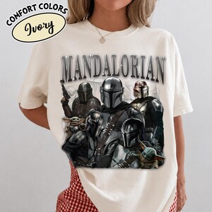 Puede incluir: Camiseta de color marfil con un gr&aacute;fico de m&uacute;ltiples personajes Mandalorianos y Grogu. La palabra "MANDALORIAN" est&aacute; impresa en la parte superior en una fuente met&aacute;lica. La camiseta tambi&eacute;n tiene las palabras "COMFORT COLORS" e "Ivory".