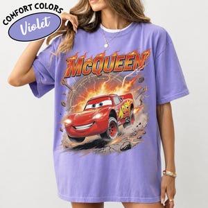 May include: A violet Comfort Colors t-shirt featuring a graphic of Lightning McQueen from the movie Cars. The design includes the name "McQueen" in fiery lettering and the number 95 on the car.