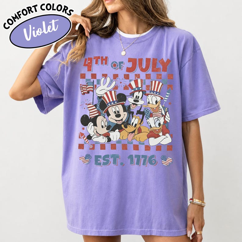 May include: A violet t-shirt featuring a graphic print of Disney characters including Mickey Mouse, Goofy, Donald Duck, and Pluto wearing patriotic hats. The text reads 4TH OF JULY and EST. 1776, with a checkered background and firework accents.
