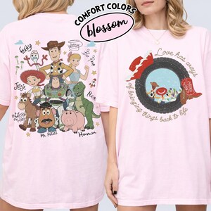 May include: A light pink t-shirt featuring Toy Story characters on the back, including Woody, Buzz Lightyear, Jessie, Bullseye, Mr. Potato Head, Rex, Hamm, Bo Peep, and Forky. The front displays a tire graphic with cowboy boots and the text Love has ways of bringing things back to life.