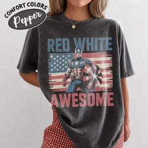 K&ouml;nnte beinhalten: Dunkelgraues T-Shirt mit einer Grafik von Captain America, seinem Schild und dem amerikanischen Flaggen-Design. Die Worte "RED WHITE AWESOME" sind auf dem Shirt aufgedruckt. Das Shirt hat die Worte "COMFORT COLORS Pepper" in einem Oval oben links.