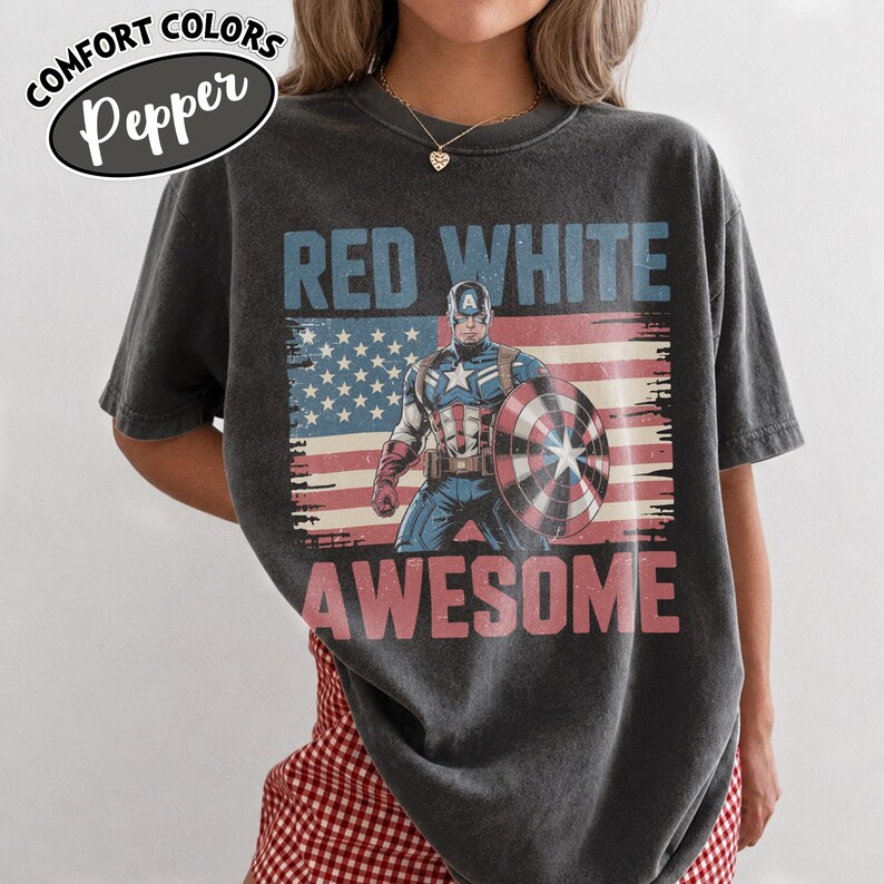 K&ouml;nnte beinhalten: Dunkelgraues T-Shirt mit einer Grafik von Captain America, seinem Schild und dem amerikanischen Flaggen-Design. Die Worte "RED WHITE AWESOME" sind auf dem Shirt aufgedruckt. Das Shirt hat die Worte "COMFORT COLORS Pepper" in einem Oval oben links.