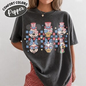 May include: Dark gray t-shirt featuring cartoon characters Mickey Mouse, Donald Duck, Minnie Mouse, Daisy Duck, Goofy, and Pluto, all wearing patriotic accessories. The shirt has a checkered background with stars and the text "COMFORT COLORS Pepper".