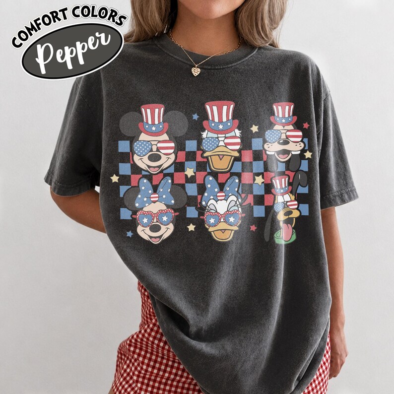 May include: Dark gray t-shirt featuring cartoon characters Mickey Mouse, Donald Duck, Minnie Mouse, Daisy Duck, Goofy, and Pluto, all wearing patriotic accessories. The shirt has a checkered background with stars and the text "COMFORT COLORS Pepper".