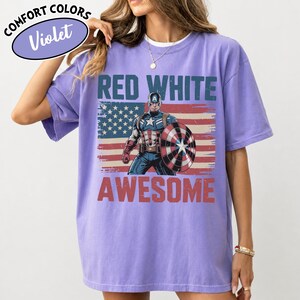 K&ouml;nnte beinhalten: Violettes Comfort Colors T-Shirt mit Captain America und dem Text "RED WHITE AWESOME" &uuml;ber einem abgenutzten amerikanischen Flaggen-Design. Das Shirt hat kurze &Auml;rmel und einen Rundhalsausschnitt.