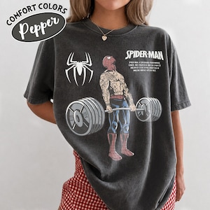 May include: Dark gray Comfort Colors t-shirt featuring a Spider-Man graphic lifting weights. The design includes a Spider-Man logo, the text "SPIDER-MAN", and the text "SPIDER-MAN A SUPERHERO FROM MARVEL COMICS WAS CREATED BY WRITER STAN LEE AND ARTIST STEVE DITKO, DEBUTING IN AMAZING FANTASY #15 IN 1962."