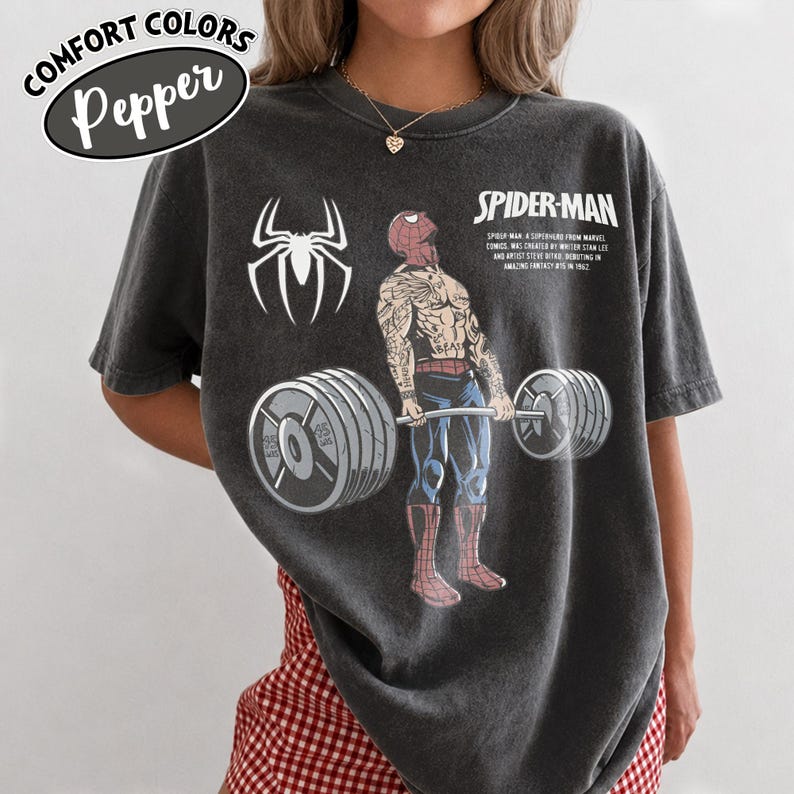 May include: Dark gray Comfort Colors t-shirt featuring a Spider-Man graphic lifting weights. The design includes a Spider-Man logo, the text "SPIDER-MAN", and the text "SPIDER-MAN A SUPERHERO FROM MARVEL COMICS WAS CREATED BY WRITER STAN LEE AND ARTIST STEVE DITKO, DEBUTING IN AMAZING FANTASY #15 IN 1962."
